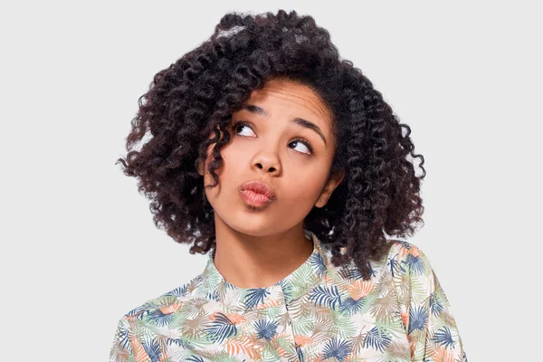 Pensando joven dama afroamericana con camisa floral, tiene expresión asombrada, mirando hacia arriba, posando sobre fondo blanco de la pared. Mujer afroamericana tiene expresión pensativa . —  Fotos de Stock