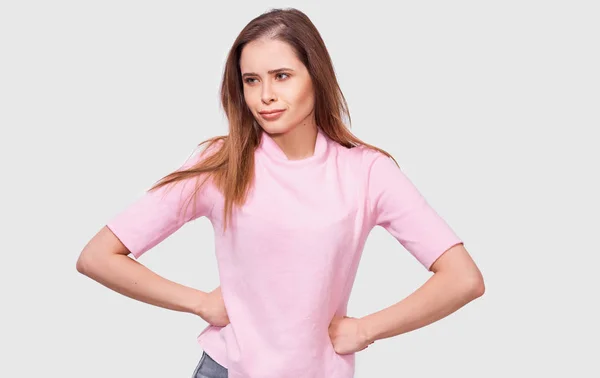 Mulher jovem tem expressão facial louco, veste roupas casuais rosa, mantém as mãos na cintura, isolado sobre fundo estúdio branco. Mulher perturbada olhando de lado, posando sobre a parede do estúdio branco . — Fotografia de Stock