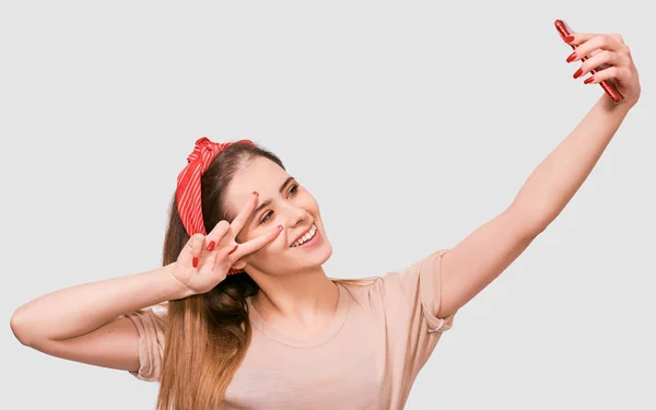 Jeune femme européenne en tenue décontractée et bandeau rouge, prenant autoportrait sur mur blanc studio. Heureuse femme souriante et prenant un selfie sur son téléphone intelligent . — Photo