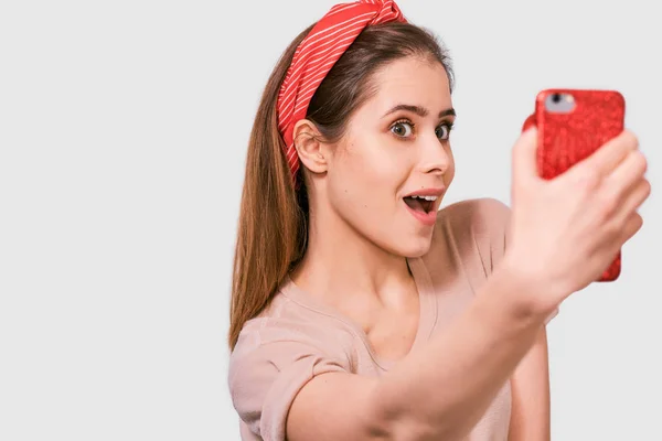 Närbild porträtt av vackra unga europeiska kvinnan i casual t-shirt och rött huvudband, tar självporträtt över vit Studio vägg. Happy förvånad kvinnlig leende och ta en selfie på hennes smarta telefon. — Stockfoto