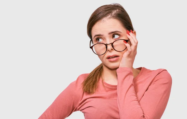 Die ernste junge Frau hat einen müden Gesichtsausdruck, trägt eine durchsichtige Brille und ein rosafarbenes lässiges Langarmshirt, posiert im Studio. horizontale Aufnahme einer ziemlich traurigen Frau, die durch Brille zur Seite schaut. — Stockfoto
