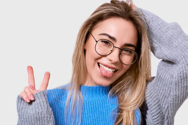 Retrato de close-up de jovem alegre mostrando língua e gesto de paz enquanto olha para a câmera e de pé contra o fundo branco. Mulher feliz vestindo roupas casuais, óculos transparentes . — Fotografia de Stock