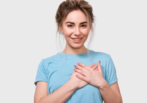 Candid portrait of pleasant young woman smiling and keeps both palms on chest, looking to the camera, dressed in blue t-shirt, posing in studio. People, emotions, feeling and body language concept — Stock Photo, Image