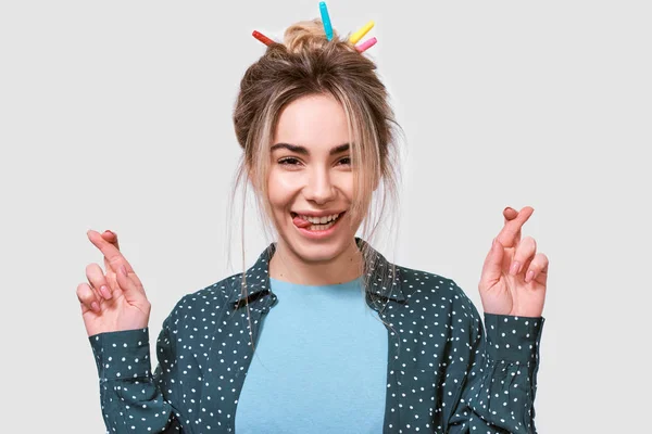 Wishful young woman smiling broadly, raises fingers crossed, makes desirable wish, wears casual outfit, posing on white wall. Happy female crosses fingers for exams. People, emotions — Stock Photo, Image