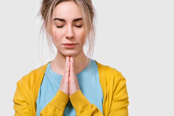 Horizontal closeup portrait of beautiful young woman praying for her family. Thoughtful female wearing yellow blouse and blue t-shirt holding hands together in praying gesture with closed eyes.