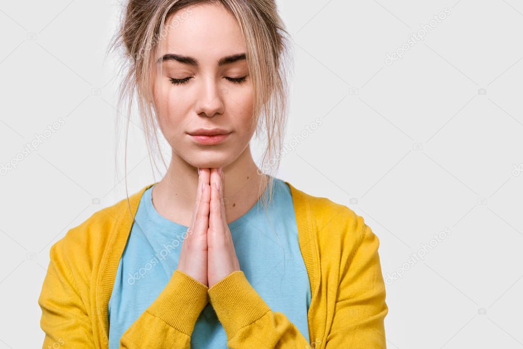 Horizontal closeup portrait of beautiful young woman praying for her family. Thoughtful female wearing yellow blouse and blue t-shirt holding hands together in praying gesture with closed eyes.