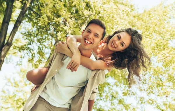 Porträtt av ung man bär sin vackra kvinna på ryggen i parken, tittar på kameran. Pojkvän ger piggyback rida till sin vackra flickvän i en sommarsemester. — Stockfoto