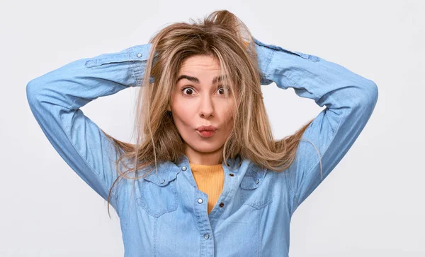 Funny young blonde woman with hands in the hair dressed casual blue shirt, making funny faces at camera, posing in studio. Positive girl having fun, making grimace — Stock Photo, Image