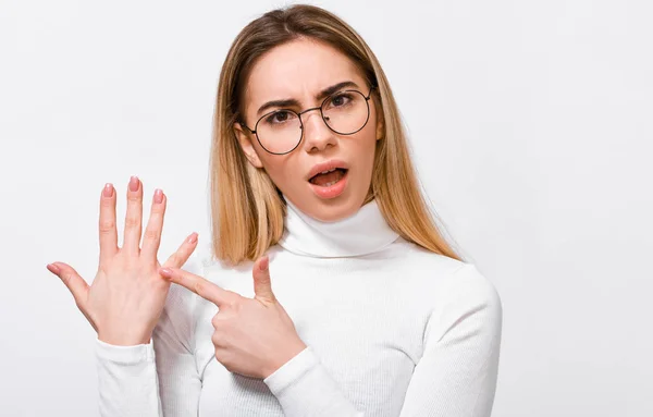 Jeune femme blonde bouleversée portant un chemisier blanc et des lunettes transparentes rondes à la mode, montrant une bague de fiançailles à son doigt, posant sur un fond de studio blanc . — Photo