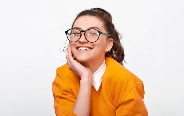 Portarit de menina estudante alegre sorrindo amplamente, usa suéter laranja e óculos redondos transparentes. Uma jovem muito positiva sente-se feliz posando sobre a parede branca do estúdio. Pessoas e emoções — Fotografia de Stock