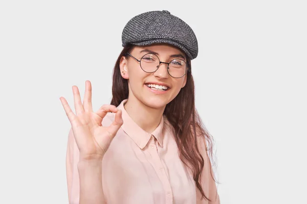 Студійний портрет позитивної молодої жінки носить модну сіру шапочку, рожеву сорочку та круглі прозорі окуляри, показуючи ОК жести. Гарненька студентка посміхається, блимає з оком і показує знак — стокове фото