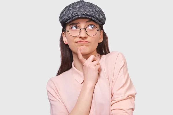 Pensive young woman wearing casual outfit and round transparent spectacles, frowning her face and  looking up to one side, poses against white studio background. People emotions concept — Stock Photo, Image