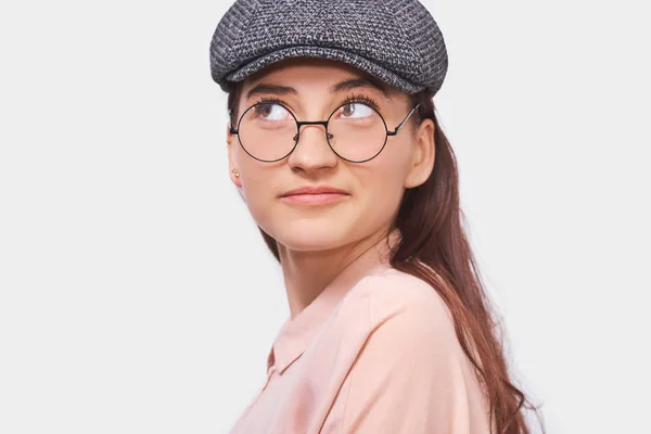 Portrait studio rapproché de jeune femme heureuse, souriante, portant une chemise rose, une casquette grise et des lunettes rondes transparentes. Jolie étudiante se sent joyeuse et posant sur le mur blanc du studio. Personnes, émotion — Photo