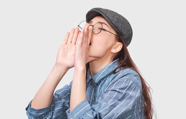 Retrato horizontal del estudio de la joven exclama en voz alta, lleva camisa azul, gorra gris y gafas redondas transparentes, aisladas sobre fondo blanco. Estudiante gritando . —  Fotos de Stock