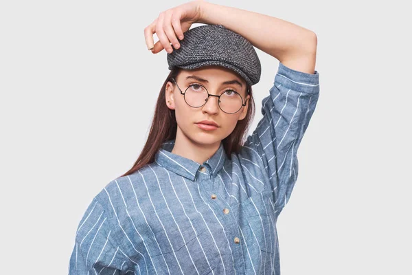 Vackra allvarliga kvinnliga bär transparenta runda Glasögon, casual blå skjorta och grå mössa, ser allvarligt direkt i kameran, poserar i studion. Människor känslor Concept — Stockfoto