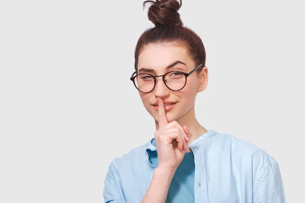 Studio close-up portrait of Caucasian pretty young woman wears transparent eyeglasses, blue shirt, holding index finger on lips, asking to keep silence. Student female asks to be quiet. — Stock Photo, Image