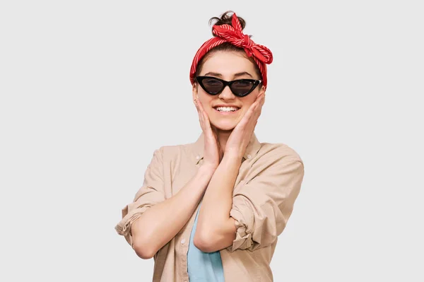 Chica estudiante encantadora sonriendo ampliamente, lleva camisa beige, diadema roja y gafas de sol negras. Mujer joven bastante positiva se siente alegre posando sobre la pared blanca del estudio. Personas y emociones —  Fotos de Stock