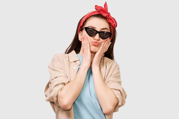 Niedliches Teenager-Mädchen bläst Luftkuss, trägt lässiges Outfit und trendige schwarze Sonnenbrille. positiver Student posiert freudig über der weißen Studiowand. Menschen und Emotionen — Stockfoto