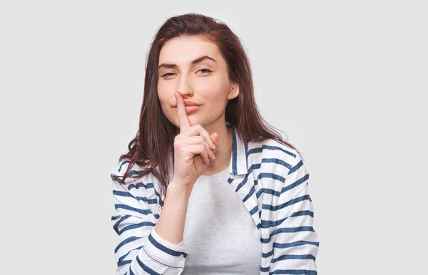 Portrait of Caucasian pretty young woman wears striped shirt, holding index finger on lips, asking to keep silence. Beautiful female asks to be quiet. People and body language — Stock Photo, Image