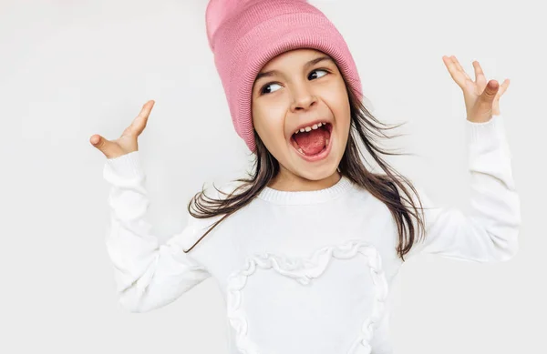 Menina feliz sorrindo amplamente com a boca aberta, olhando para um lado, vestindo suéter branco e chapéu rosa de malha, as mãos se levantam, posando sobre a parede cinza do estúdio. Criança bonito à espera de Natal . — Fotografia de Stock