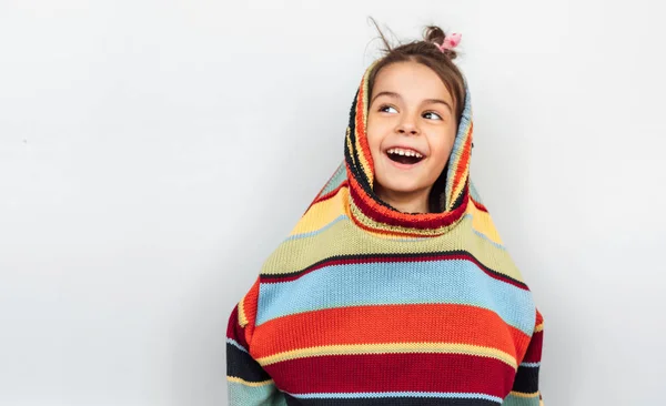 회색 배경에 큰 화려한 스웨터와 함께 연주, 광범위하게 미소 행복 한 어린 소녀의 클로즈업 초상화. 하얀 벽 너머의 빈 복사 공간을 올려다보며 웃는 아이가 스웨터를 들여다봅끈습니다. — 스톡 사진