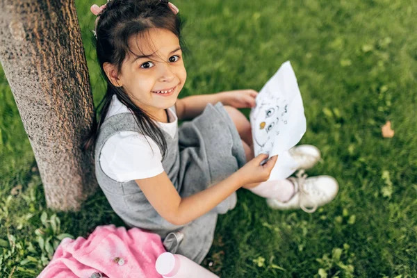 Sevimli küçük kız gülümseyen ve kağıt üzerinde bir şey öğrenme, yeşil çim ağacın altında oturan, sırt çantası dışında üst görünümü. Güzel çocuk öğrenci okul gününden sonra rahatlatıcı. Eğitim