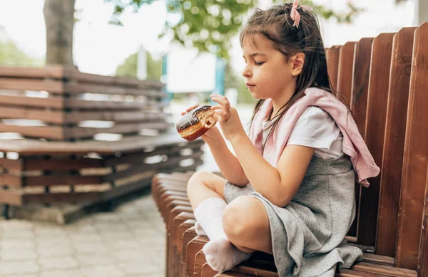 屋外のベンチに座ってドーナツを食べるかわいい女の子の学生。愛らしい子供の生徒は、学校の休憩中に軽食の時間を持っています。不健康な食べ物子供のためのおいしい食べ物。教育と健康の概念. — ストック写真