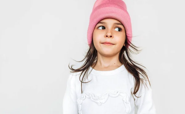 Estúdio close-up retrato da menina agradada sorrindo e olhando para um lado, vestindo suéter branco e chapéu rosa de malha, posando sobre a parede cinza claro. Bela criança esperando por um tempo de Natal . — Fotografia de Stock