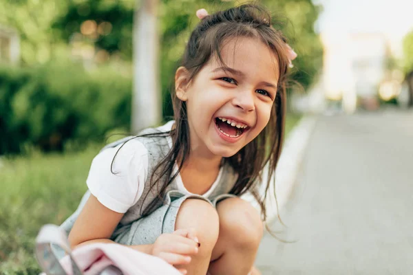 Close-up outdoors portrait of happy cute little girl laughing and sitting on doorstep with backpack after school. Happy child pupil relaxing on the street after kindergarten. People, education concept — Stock Photo, Image