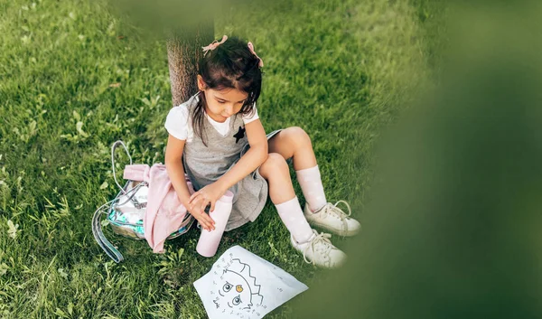 Kültéri kép aranyos kislány ül a fa alatt a zöld fű, a hátizsák és a palack a víz, ami a papíron. Gyönyörű gyermek tanuló pihentető iskola után nap. Oktatási koncepció — Stock Fotó