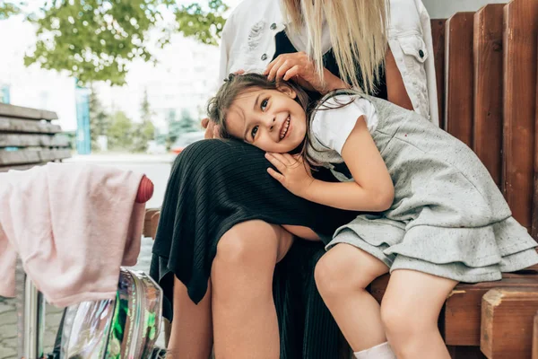 Imagen horizontal de la niña linda y feliz relajándose en las piernas de la madre abrazándola antes del día del jardín de infantes. Mamá con niño en edad preescolar sentado en el banco en la calle de la ciudad . — Foto de Stock