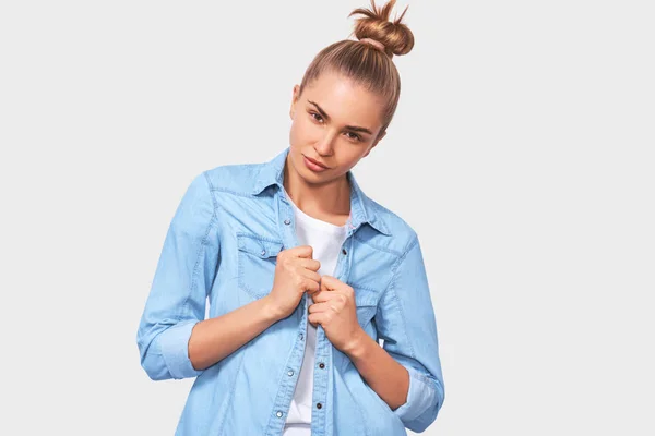 Estudante estudante com penteado de coque olhando seriamente de pé sobre fundo estúdio branco. Jovem mulher vestindo camisa jeans azul posando no fundo do estúdio . — Fotografia de Stock