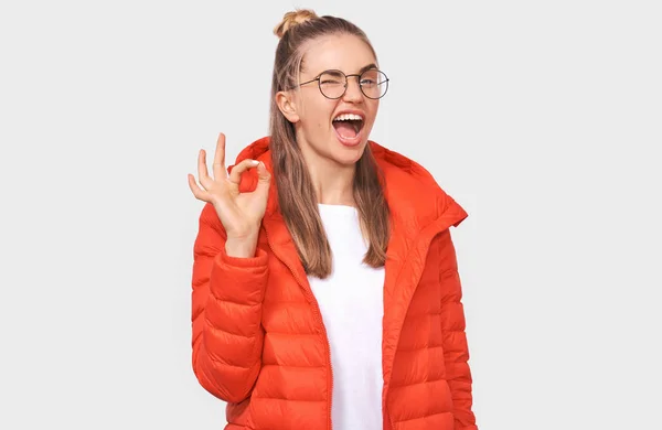 Portrait of cheerful young woman wearing red jacket, transparetn eyeglasses, showing Ok gestures with hand. Pretty female smiling broadly, posing over white background. — Stock Photo, Image