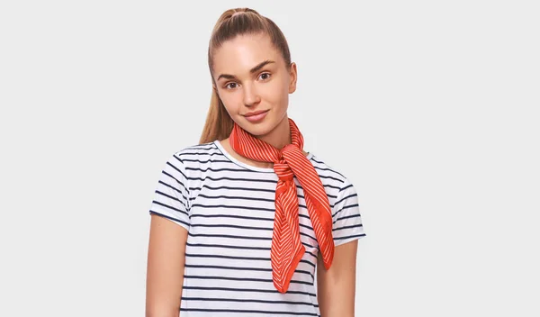Europeisk charmig ung kvinna med hästsvans frisyr, klädd randig t-shirt, snygg röd halsduk på halsen, ser till kameran poserar över vit Studio bakgrund. — Stockfoto