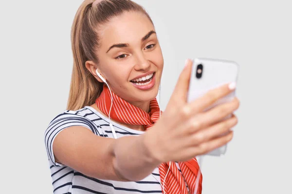 Belle jeune femme prenant autoportrait, portant un t-shirt rayé, écharpe rouge sur le cou en studio. Happy femelle faire un selfie sur son téléphone intelligent ou avoir une conversation avec son ami en ligne . — Photo