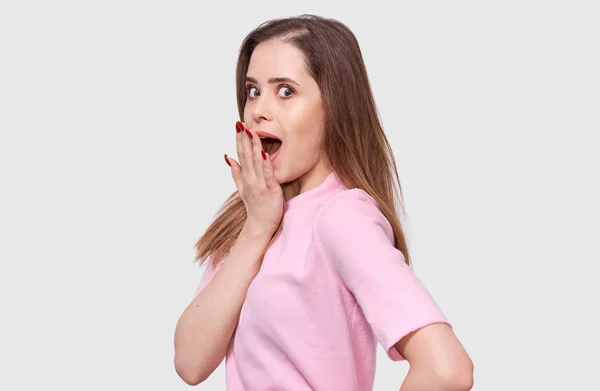 Shoked young woman has amazed expression, wearing pink shirt. Portrait of pretty amazed female, holding hand on mouth with red nails, looking astonished posing over white background. People emotions — Stock Photo, Image