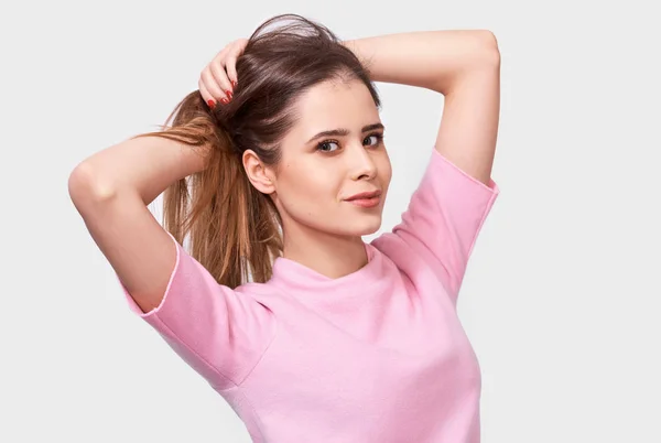 Porträtt av attraktiv ung kvinna leende, klädd i rosa t-shirt, poserar mot vit bakgrund. Pretty hona har glädjande uttryck, vilket gör hästsvans frisyr, poser i studion. — Stockfoto