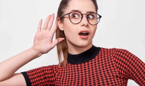 Zdumiona młoda kobieta zwracając uwagę i umieszczając rękę na uchu prosząc kogoś, aby mówić głośniej lub szemranie, izolowane na białej ścianie. Ładna dziewczyna, która słyszy tajne rozmowy, noszenie okularów. — Zdjęcie stockowe