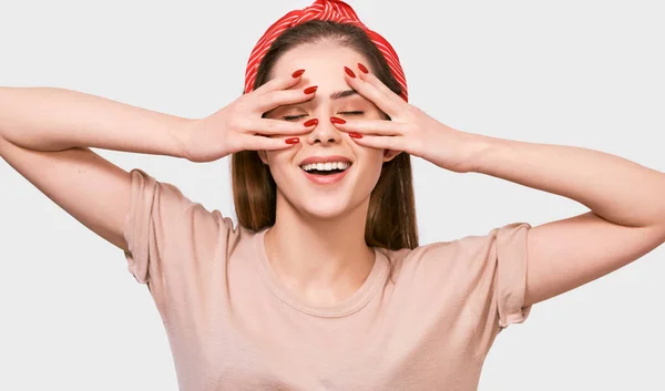 Glückliche junge Frau, die ihr Gesicht mit den Fingern verbarg und ihr Auge verdeckte. Hübsche Frau, die ihr Gesicht mit der Hand bedeckt, trägt beiges T-Shirt, rotes Stirnband und rote Nägel, mit geschlossenen Augen posiert an weißer Wand — Stockfoto