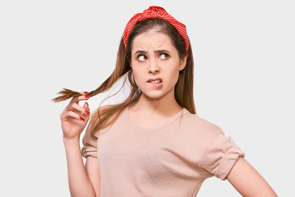 Студійний портрет вдумливої красивої молодої жінки носить бежеву футболку і червоний головний убір, який грає з шматочком волосся, виглядає невпевнено, позує на білому тлі. Люди, концепція емоцій — стокове фото