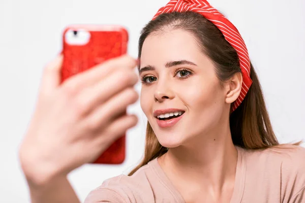 Closeup portrait of beautiful young brunette woman in casual clothes and red headband, taking self portrait over white studio wall. Happy female smiling and taking a selfie on her smart phone. — Stock Photo, Image