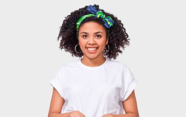 Beyaz tişört giymiş, mutlu hisseden güzel, koyu tenli genç bir kadın. Afro-Amerikan kadın geniş gülümseyerek, beyaz t-shirt ve trandy kafa bandı beyaz stüdyo duvar üzerinde poz giyiyor