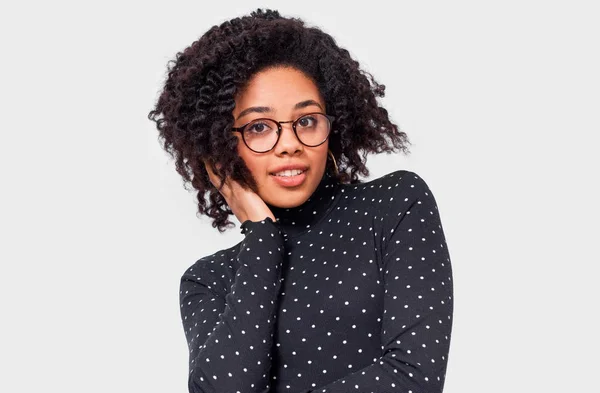 Mujer joven de piel oscura bastante soñadora vestida de negro con blusa de puntos blancos, cogida de la mano en la cabeza, se siente feliz. Mujer afroamericana sonriendo ampliamente, usando gafas redondas posando sobre pared blanca —  Fotos de Stock