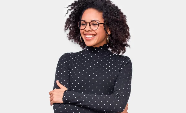 Jovem feliz vestida de preto com blusa branca, segurando as mãos cruzadas, sinta-se feliz. Afro-americano feminino sorrindo amplamente, vestindo óculos redondos transparentes posando sobre a parede branca — Fotografia de Stock