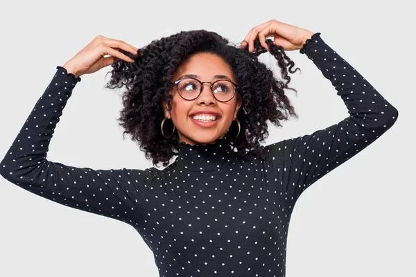 Uma jovem de pele escura vestida de preto com blusa branca, de mãos dadas no cabelo, feliz. Afro-americano feminino sorrindo amplamente, vestindo óculos redondos posando em estúdio — Fotografia de Stock