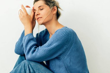 Sevimli genç kadın elinde kapuçino bir kupa ile rahat örme mavi kazak ve kot giyiyor. Güzel genç kadın evde sıcak içecek içiyor. Bir kahve fincanı tutan lovely sarışın kadın 