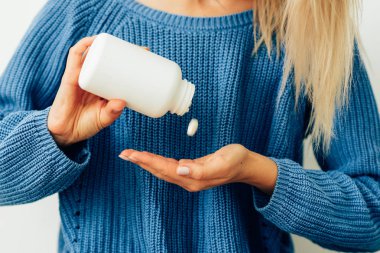 Kadının elinin yakın çekim kırpılmış görüntüsü hapları şişeden dışarı döküyor. Mavi örme kazak giyen genç sarışın kadın beyaz bir şişeden eline bazı haplar döküyor. Sağlık ve tıp.