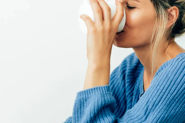 Retrato de close-up de jovem feliz bebendo bebida quente em casa. Mulher loira sorridente segurando uma xícara de café. Mulher caucasiana vestindo camisola azul e sentada em casa com xícara de chá em suas mãos . — Fotografia de Stock