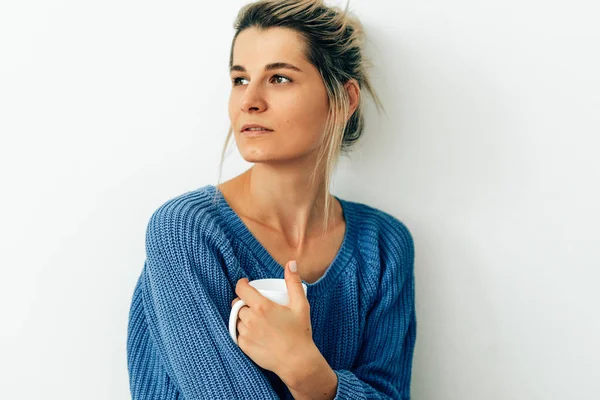 Imagem espontânea de mulher atraente vestida com camisola azul de malha aconchegante com uma caneca de cappuccino. Mulher bonita bebendo bebida quente em casa. Mulher loira encantadora segurando uma xícara de café . — Fotografia de Stock