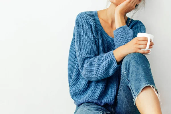 Cropped image of pretty young woman wearing cozy knitted blue sweater and jeans with a mug in her hand. Beautiful woman drinking hot beverage at home. Lovely blonde female holding a coffee cup. — Stock Photo, Image